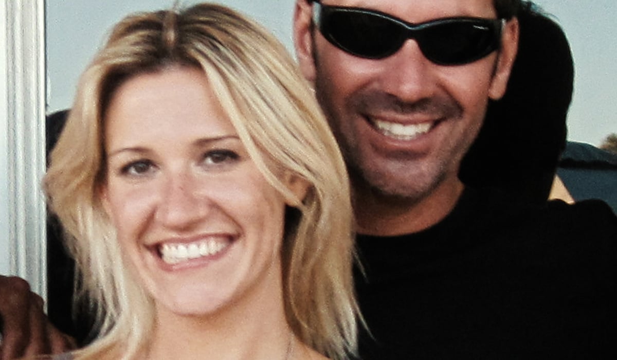 "Smiling woman with blonde hair and a man in sunglasses standing close together outdoors, both appearing happy with sunlight and greenery in the background."