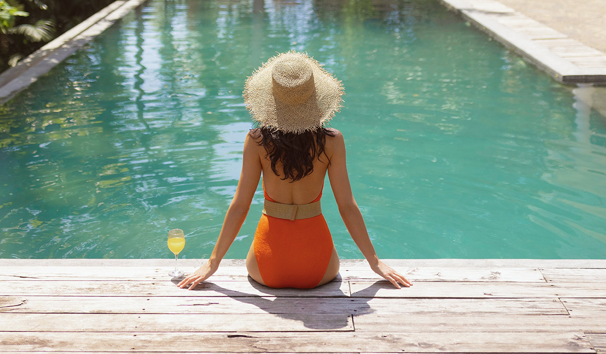 Rear view of stylish woman in orange swimsuit and straw hat with cocktail by swimming pool. Tropical holiday and spa lifestyle concept, ideal for luxury travel promotions, wellness, and leisure ads.; Shutterstock ID 2727035877; purchase_order: -; job: -; client: -; other: