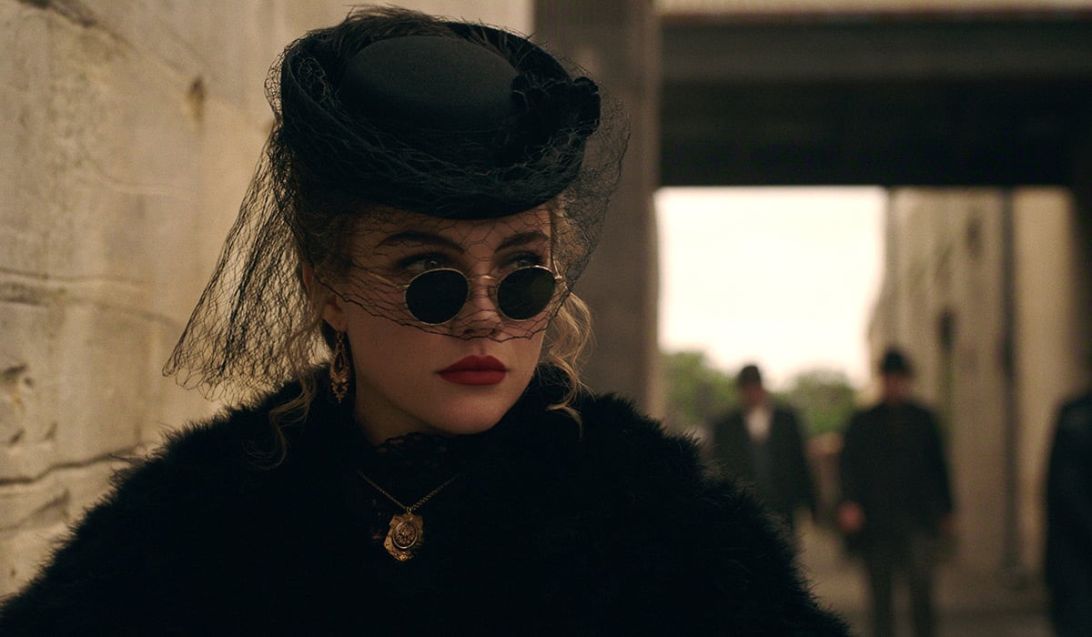"Woman in vintage black attire with round sunglasses, a veiled hat, and gold jewelry stands near a stone wall in an urban setting, with blurred people and old-style architecture in the background."